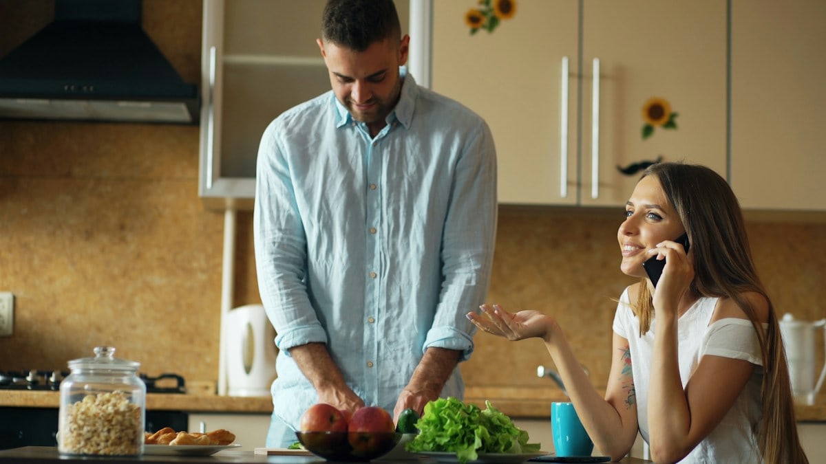 Attractive couple in the kitchen early morning. Beautiful girl chatting phone while her boyfriend cooking breakfast at home